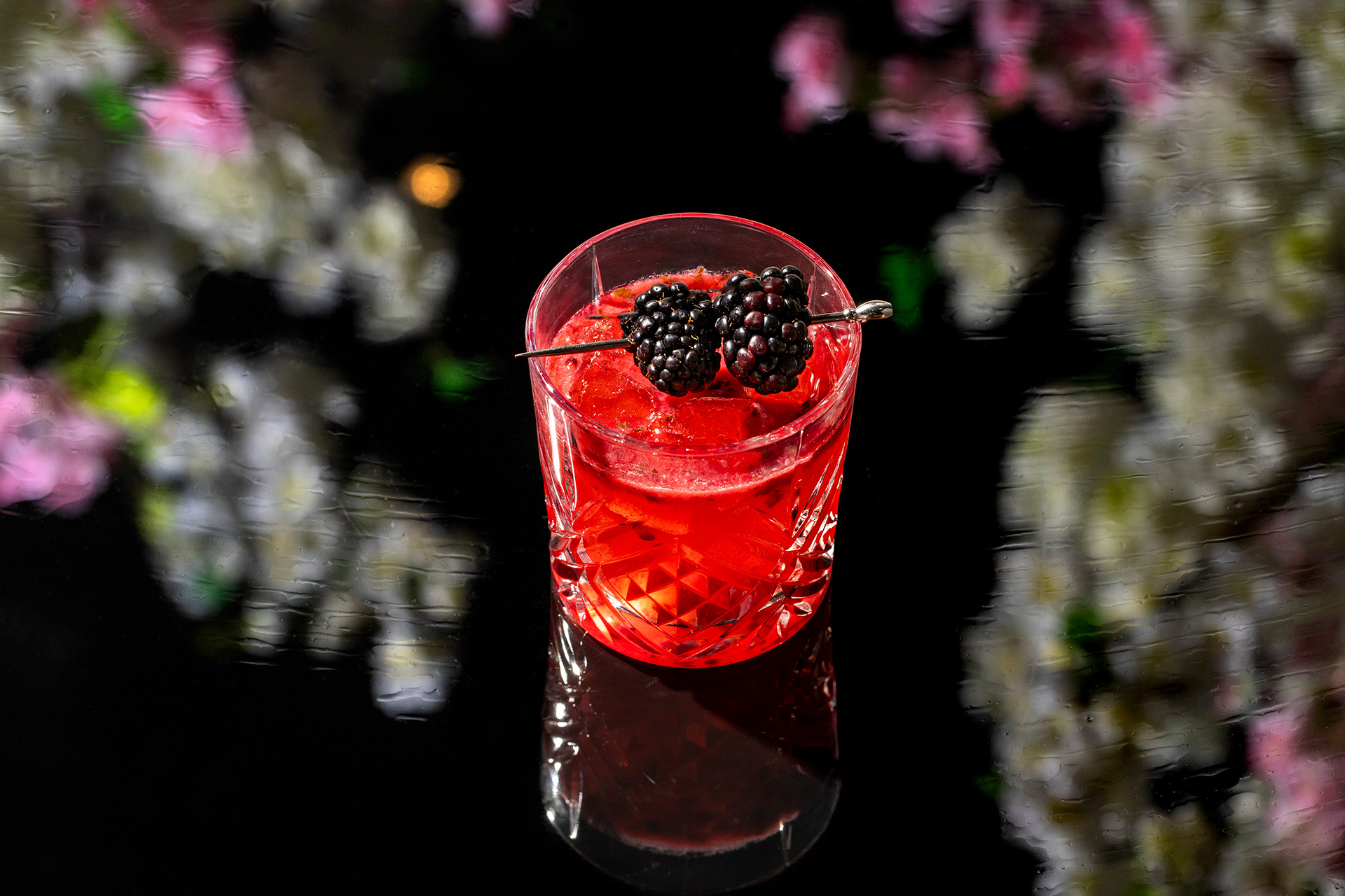 A crystal glass filled with a ruby-red cocktail, garnished with a blackberry skewer, floral bokeh softly lit behind
