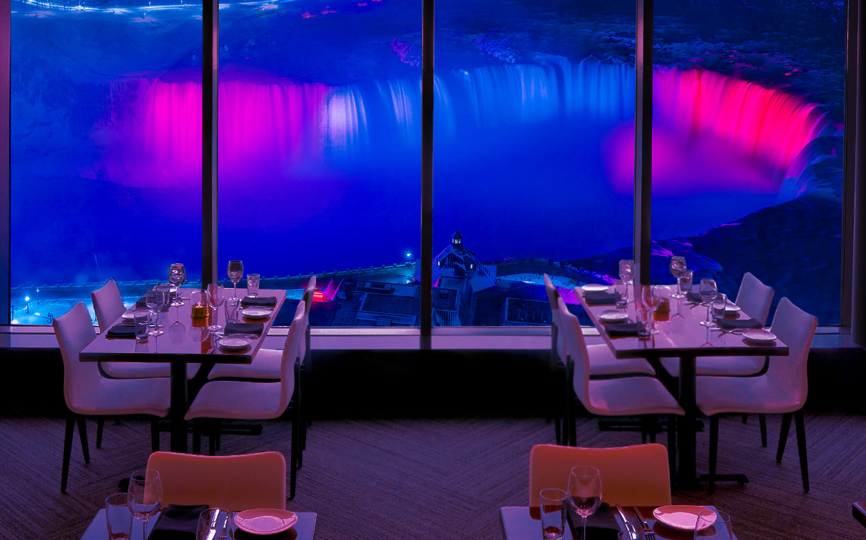 The Horseshoe Falls illuminated in blue and pink at night, viewed through the panoramic windows of the STK Niagara Falls dining room