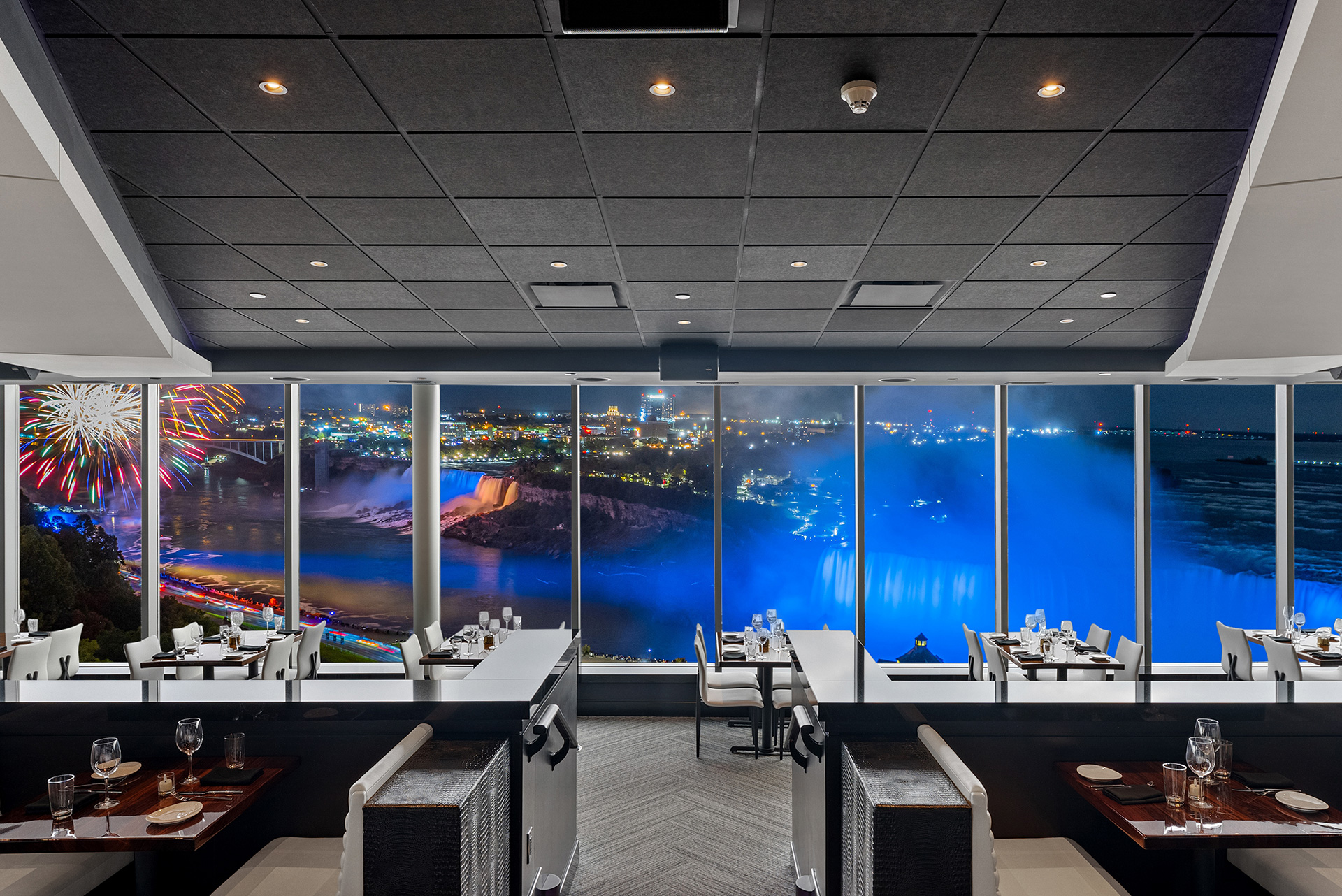 The STK Niagara Falls dining room at night with fireworks bursting above the American Falls, seen through the panoramic windows on the 9th floor