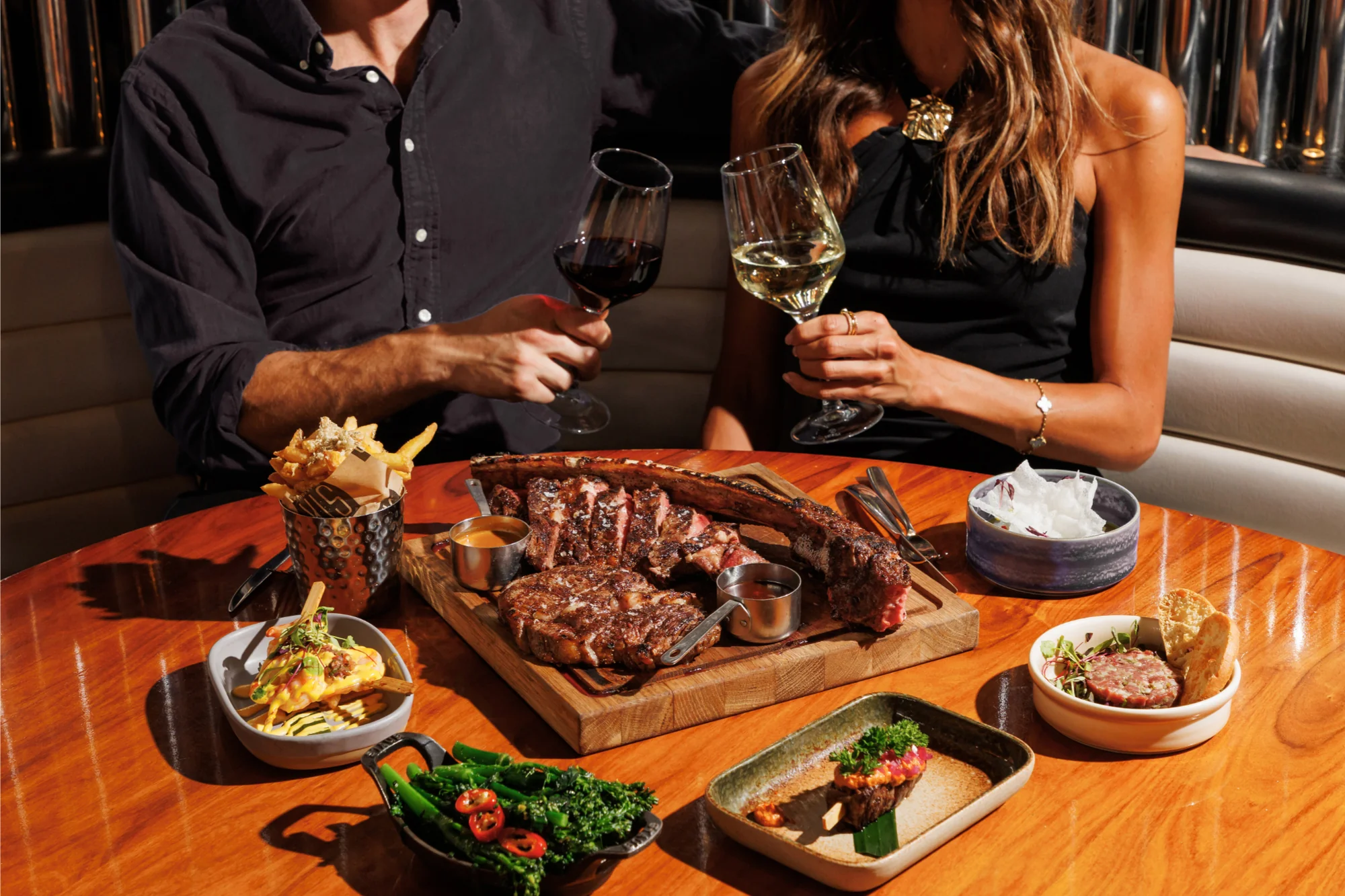 A couple toasting red and white wine over a Tomahawk steak, fries, broccolini, tartare, and shared plates at STK Niagara Falls