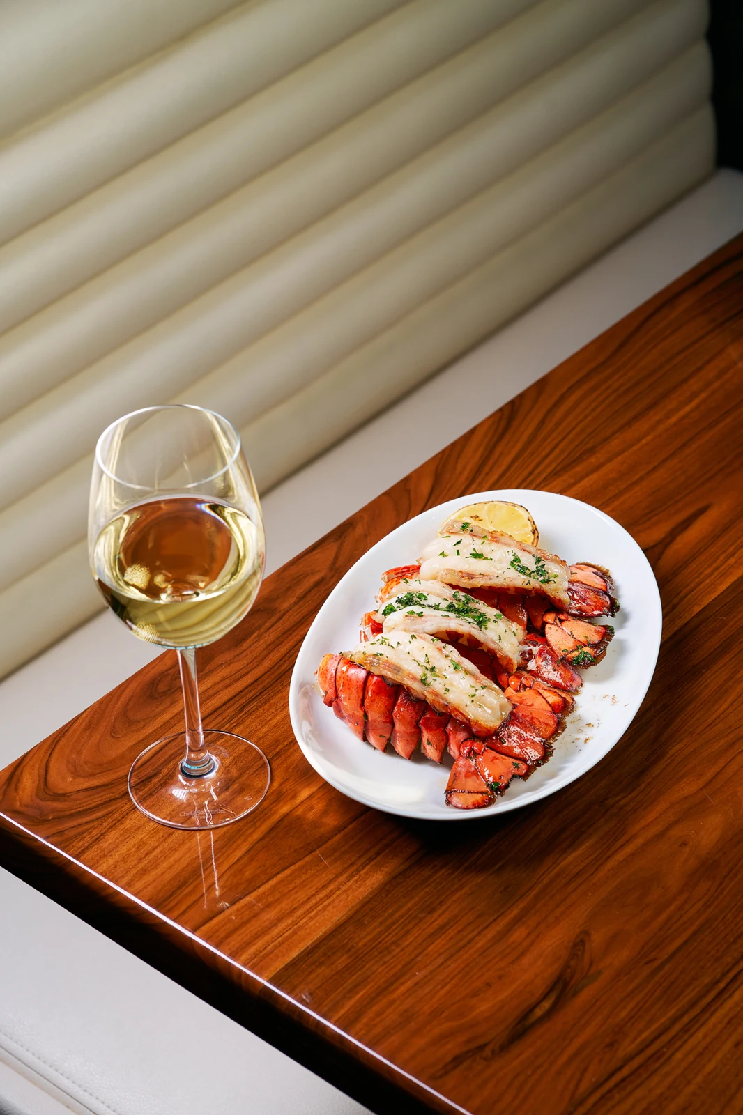 A butter-basted lobster tail plated on white ceramic beside a chilled glass of white wine on a walnut banquette table at STK Niagara Falls