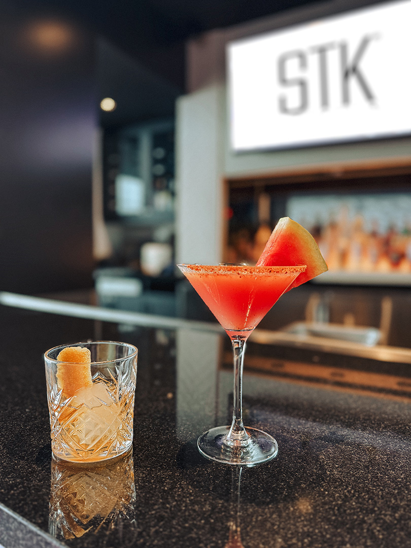 A watermelon martini and a spiced old fashioned resting on the STK Niagara Falls bar counter, the illuminated STK sign glowing behind