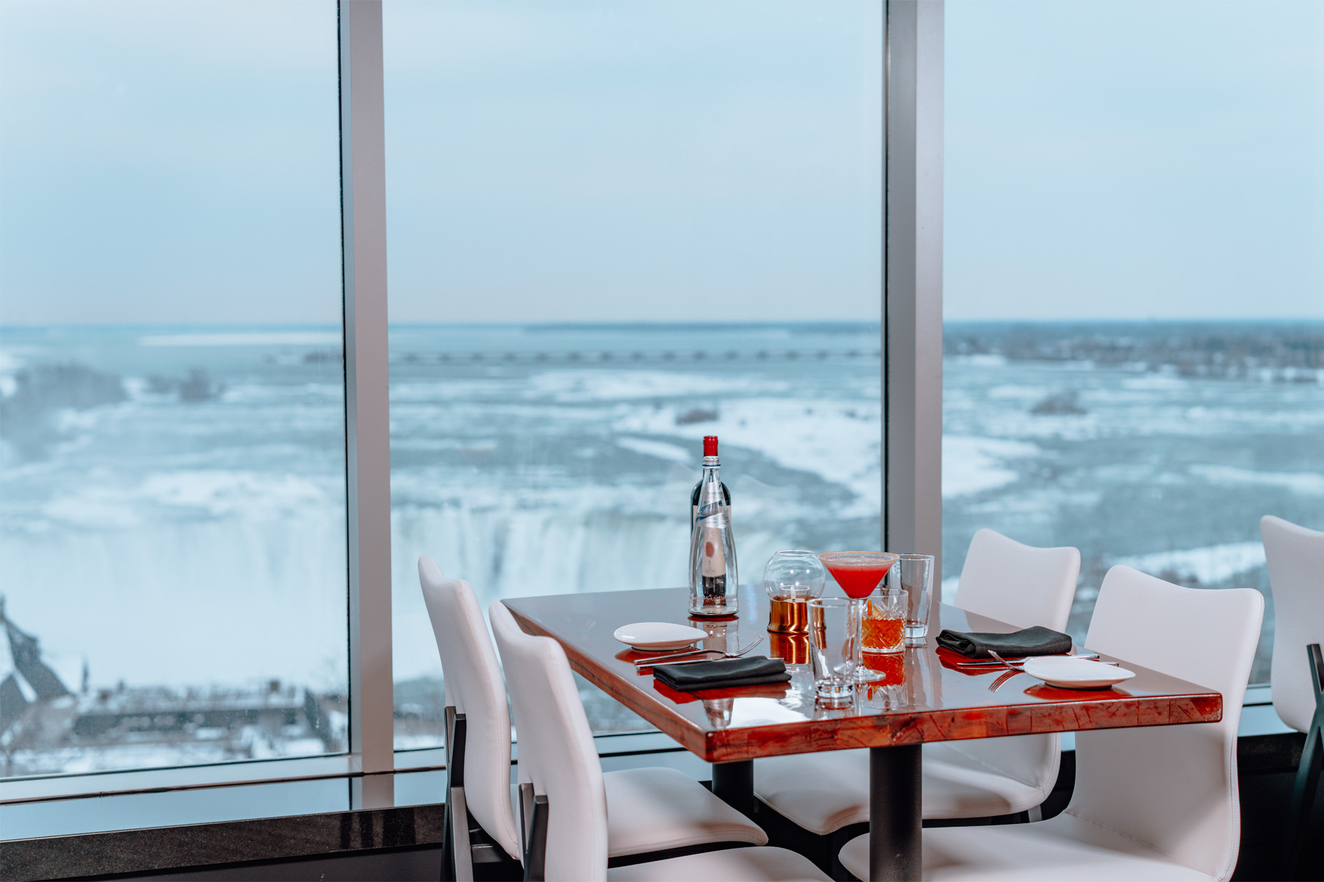 A window-side table at STK Niagara Falls set with stemware and a cocktail, the Horseshoe Falls in soft winter light beyond the panoramic window