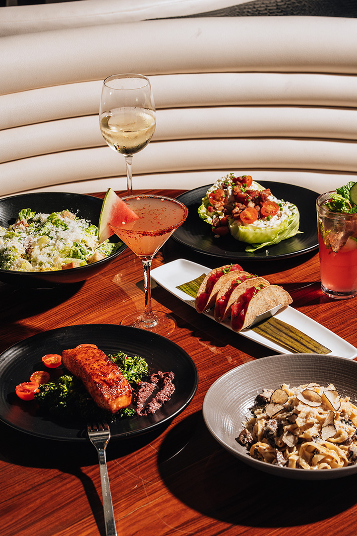 An STK Niagara Falls tabletop spread with pan-seared salmon, Ahi tuna tacos, a wedge and Caesar salad, truffle pasta, and cocktails set on a walnut booth table