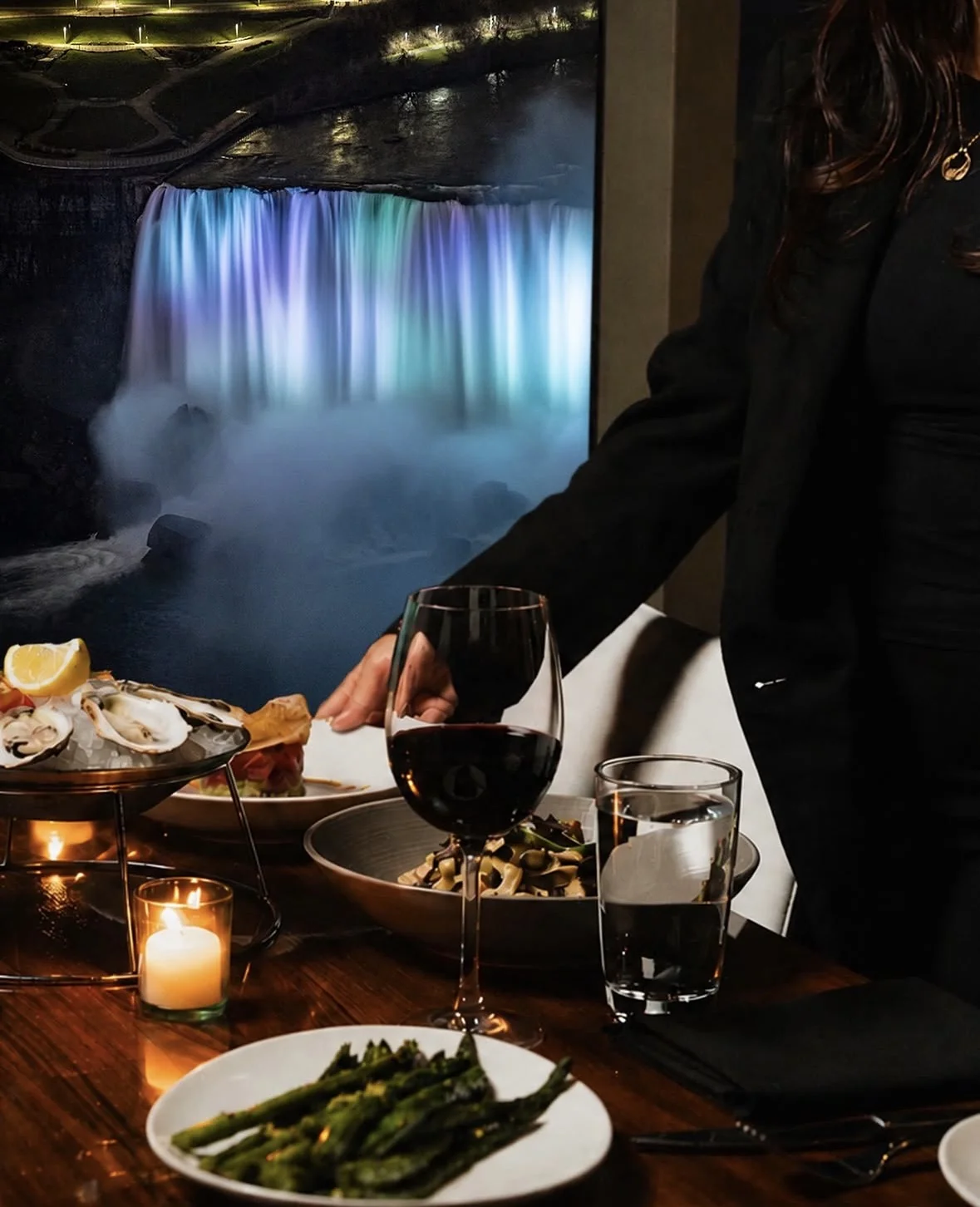 A server presenting wine, oysters, truffle pasta, and grilled asparagus beside the illuminated Falls art panel at STK Niagara Falls
